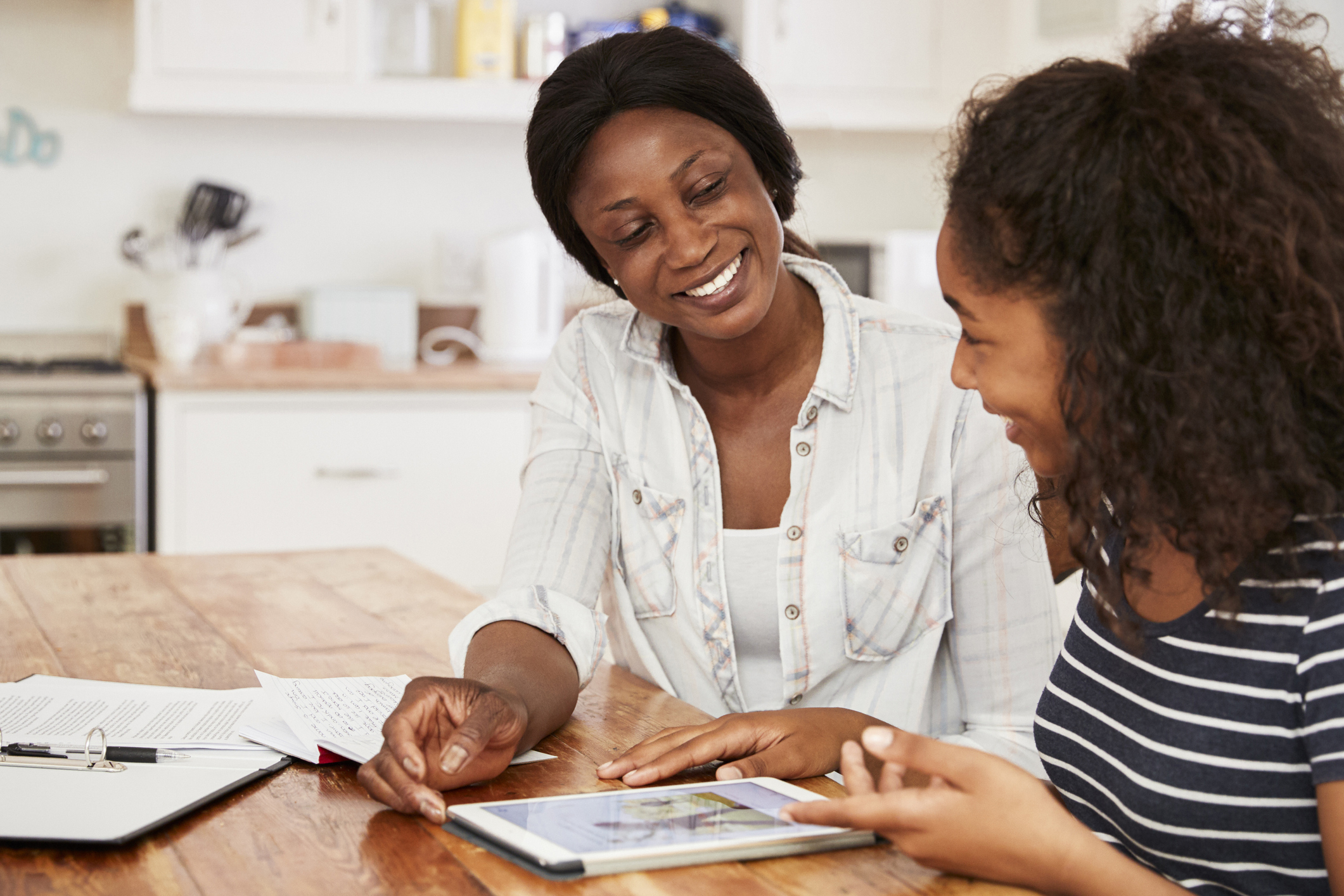 A mother and daughter sitting a a table in the kitchen. They are looking at a tablet computer.