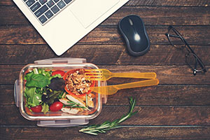 A salad sits on a desk next to a laptop computer.