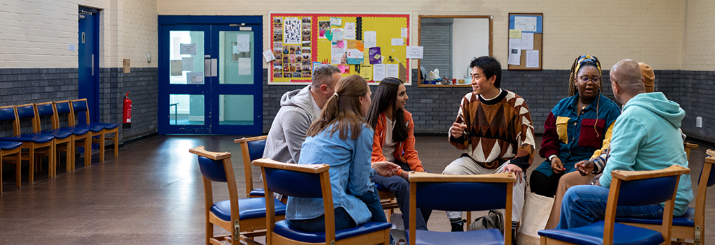 A multi racial group of people meeting; they sit in blue chairs arranged in a circle.