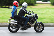 Two riders on a silver motorcycle. Each rider is wearing a full-face helmet