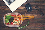 A salad sits on a desk next to a laptop computer.